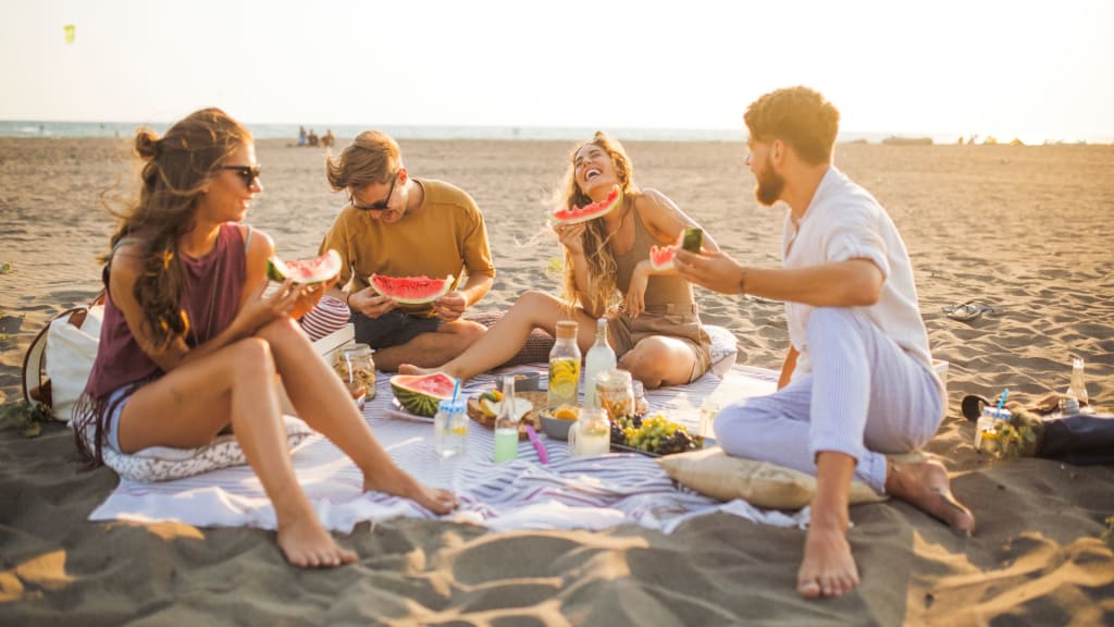 Four Americans living in Costa Rica enjoy a beach picnic, savoring watermelon and other treats on a blanket with the serene sea as their backdrop.